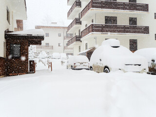 Snowy road with cars covered with snow on the background of cottages. Snowfall in the ski resort