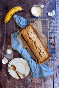 Flat lay with banana bread on a cooling rack with eggs, bananas, walnuts, flour and plates on wooden background
