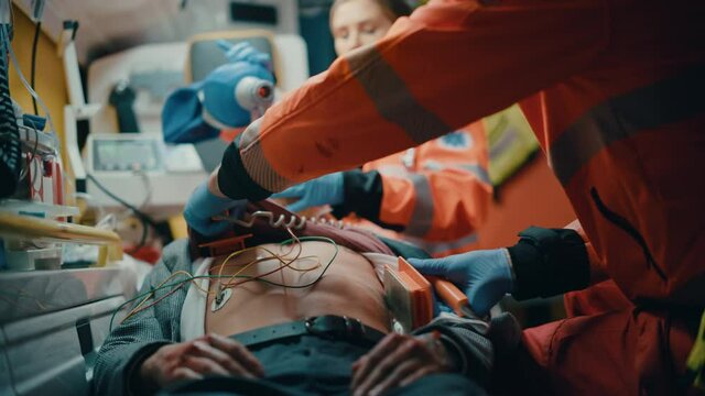 EMS Paramedics Team Provide Medical Help to Injured Patient on the Way to Hospital. Emergency Care Assistants Using a Defibrillator and Ventilation Mask to Bring the Man Back to Life in an Ambulance.