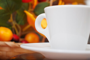 White porcelain conical cup with an eyelet on a saucer close-up. A cup on the background of a basket with a bouquet of imitation of branches of autumn trees with yellowed leaves.