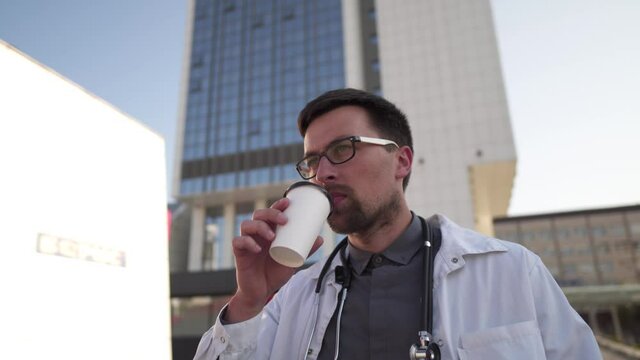 Medical professional during coffee break after long day. General practitioner drinking coffee to go while walking on the background of the clinic. Doctor in a lab coat drinks a hot drink