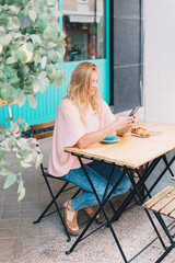 Attractive blonde woman looking at her cell phone on a cafeteria terrace while eating breakfast.
