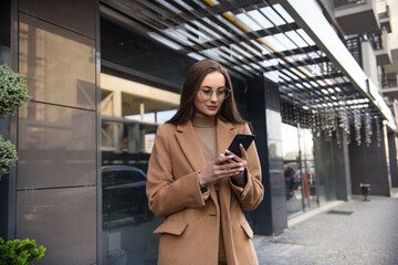 Business woman curly woman wearing trendy sunglasses walks down the central city street and uses her phone. Pretty summer woman in brown jacket walks down the street looking at her mobile phone.