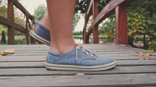 Female Feet wearing Denim Sneakers Shoes crossing Wooden Bridge in the Autumn Park, Close Up on boots. 4K. Unrecognizable young woman spending time in the evening fall garden. Steadicam Dolly Shot