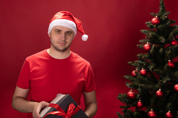 thoughtful Unshaven young man in Santa Claus hat and red T-shirt holds gift box with Christmas...