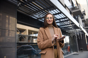 Smiling curly businesswoman wearing trendy sunglasses walks down the central city street and uses her phone. Businesswoman holding cell phone.
