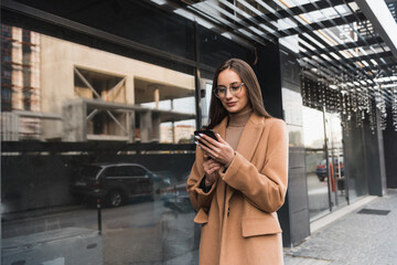 An attractive young woman walking in the city center, texting and laughing. Beautiful woman in eyewear and beige coat using smartphone while standing near dark building.