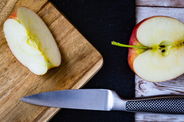 Red gala apple cut in half and a quarter slice of the same apple placed on wooden cutting board over black stone background on wooden table next to kitchen knife