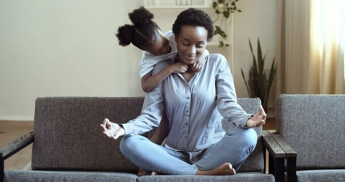 Active preschool african daughter tickles ethnicity mother sitting in lotus position practice self-control feel serenity calmness, reduce stress fatigue during day at home with little mixed race child