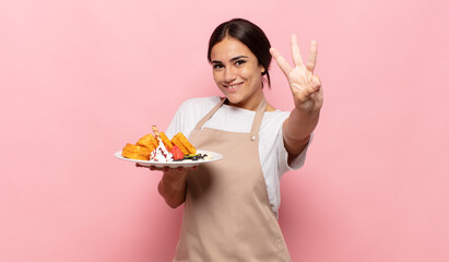 young hispanic woman smiling and looking friendly, showing number three or third with hand forward,...