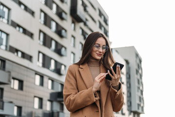 Business woman in autumn coat browsing internet on smartphone outdoors. Smiling lady with long brown hair using modern gadgets on street.