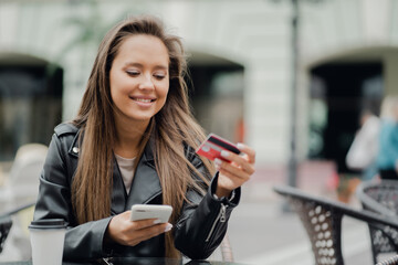 shopping online store, a young woman makes purchases through a mobile phone device. Sitting in a cafe on the street and smiling, good mood