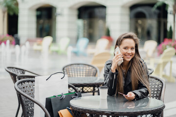 portrait of a Thrifty confident brunette woman of European appearance holding a Bank card and phone calling a friend, sitting in a street cafe and drinking coffee to take away.