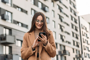 Happy pretty brunette girl using mobile phone near office, beautiful woman browsing phone and doing...
