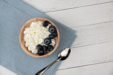 wooden bowl of farmer cheese and blueberry and a spoon on blue napkin on white wooden background flat lay. Healthy eating concept