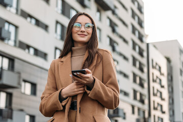 Happy pretty brunette girl using mobile phone near office, beautiful woman browsing phone and doing online shopping in a mobile app while smiling walking near shopping mall.