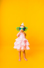 a little girl in a pink puffy dress and a paper hat and big funny glasses blows a whistle on a yellow background
