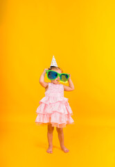 A little girl in a pink dress hides behind big funny glasses on a yellow background