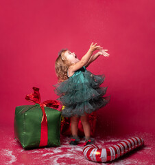 Toddler girl in green Christmas dress with Xmas candy cane and gifts on a red background.