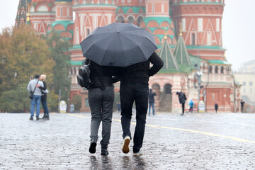 Rain in a Moscow, couple in autumn clothes with one umbrella walking on the Red Square on background of St. Basil's Cathedral. People in rainy weather, fall season