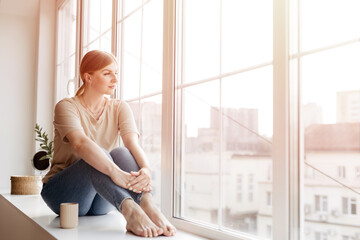 Attractive young woman sitting on the windowsill and looking out of the window daydreaming