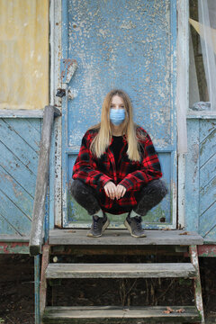 A young blonde woman in a red shirt and a medical mask on her face is squatting on the threshold of an old wooden house.
