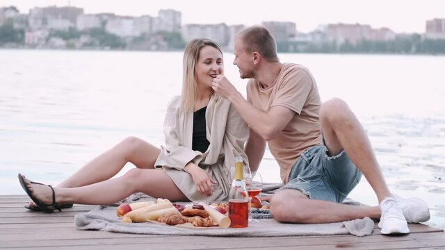 Loving couple are on a date. They are having a picnic at the lake. The man and woman are sitting on a wooden berth with lots of food on a blanket. Wedding proposal concept.