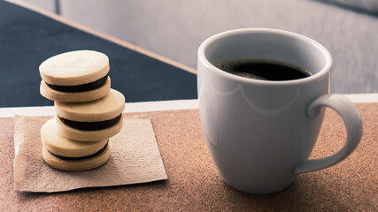 Primer plano frontal de una taza de cerámica blanca de café negro caliente junto a tres pequeños alfajores rellenos de dulce de leche hechos y horneados en casa. 