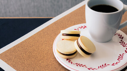 Primer plano desde un ángulo levemente alto de una taza de café negro de cerámica blanca junto a tres pequeños alfajores rellenos de dulce leche horneados en casa.