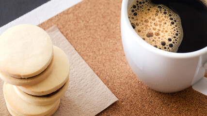 Primer plano desde un ángulo cenital de una taza de cerámica blanca de café negro caliente junto a tres pequeños alfajores rellenos de dulce leche horneados en casa y bajo iluminación natural. 