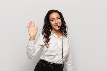 young hispanic woman smiling happily and cheerfully, waving hand, welcoming and greeting you, or...