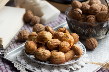 Cookies in the form of walnuts on a metal tray on a checkered tablecloth, next to them are real nuts in shells, an open book and other decor.