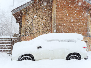 A snow-covered car is parked near a wooden hut on a cloudy day with heavy snow