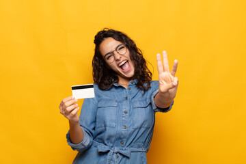 young hispanic woman smiling and looking friendly, showing number three or third with hand forward,...