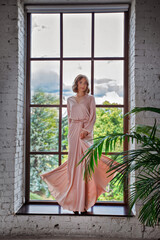 slender young woman in a beautiful pink long dress  stands at the big window