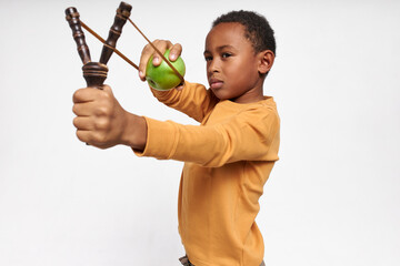 Isolated image of serious concentrated little black boy holding Y-shaped stick with elastic, shooting green apple, having focused facial expression. Accurate African child playing with slingshot - obrazy, fototapety, plakaty
