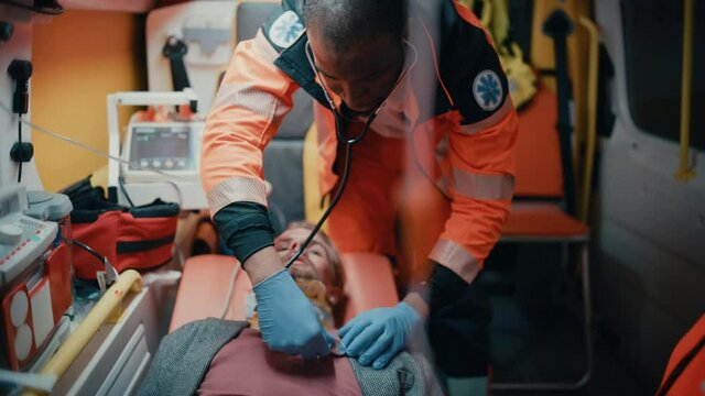 Female and Male EMS Paramedics Ride to Healthcare Hospital with an Injured Patient on the Ambulance. Black African American Emergency Care Assistant Checking Up on the Sick Young Man with Stethoscope.