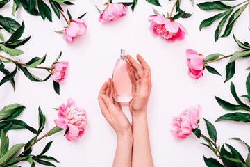 Pink bottle of women's perfume in female hands and peony flowers around on white background. Spring...