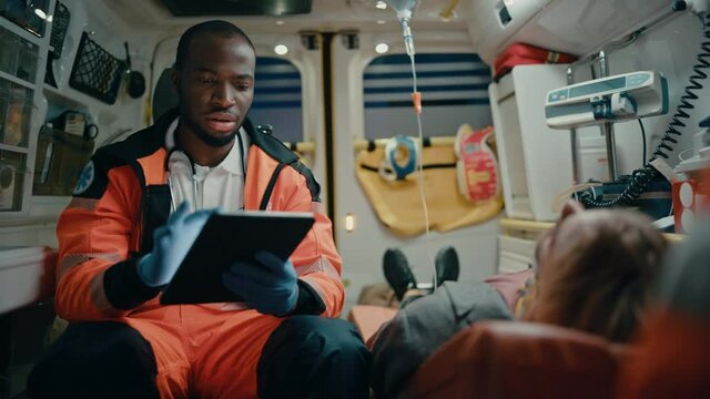 Black African American EMS Professional Paramedic Using Tablet Computer to Fill a Questionnaire for the Injured Patient on the Way to Hospital. Emergency Medical Care Assistant Works in an Ambulance.