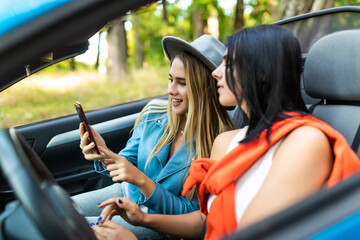 Fototapeta na wymiar Young two women sitting in convertible car and use phone serfing in social networks.