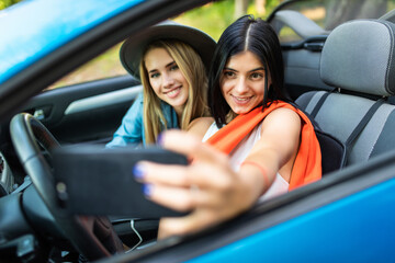 Two cheerful young women sitting on car and taking selfie with phone