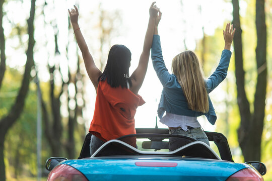 Two excited women stand in the back of open car. Back view