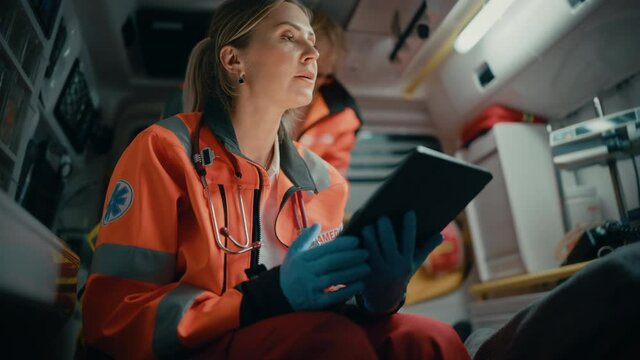 Female EMS Professional Paramedic Using Tablet Computer to Fill a Questionnaire for the Injured Patient on the Way to Hospital. Emergency Care Assistant Comforting the Patient in an Ambulance.