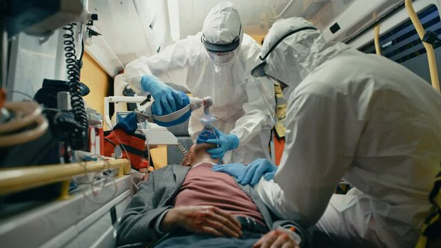 Team of EMS Paramedics in Disposable Coverall Suits Provide Medical Help to Injured Patient on the Way to Hospital. Emergency Care Assistants Putting On Non-Invasive Ventilation Mask in an Ambulance.
