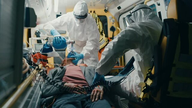 Team of EMS Paramedics in Disposable Coverall Suits Provide Medical Help to Injured Patient on the Way to Hospital. Emergency Care Assistants Putting On Non-Invasive Ventilation Mask in an Ambulance.