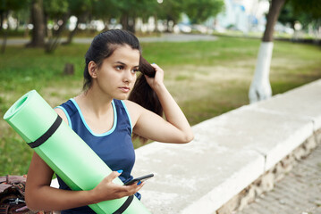 Close-up of young sporty woman in blue sports clothing holding exercise mat and looking at her mobile phone in her hand while standing outdoors