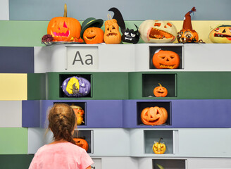 Young girl looking at the wall with various halloween carved orange pumpkins crafted by children at the education school, rear view.