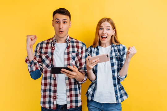 happy couple, a man and a woman, play video games on a smartphone, rejoice and show a winning gesture, on an yellow background