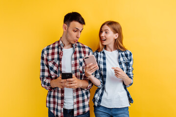 shocked young couple, man and a woman in plaid shirts, holding mobile phones, on a yellow background