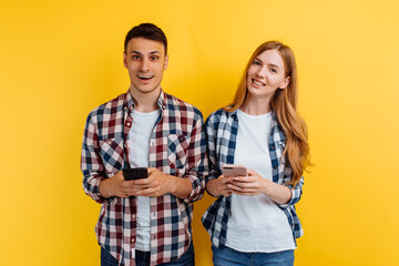 young couple, a man and a woman in plaid shirts, holding mobile phones in their hands, on a yellow background